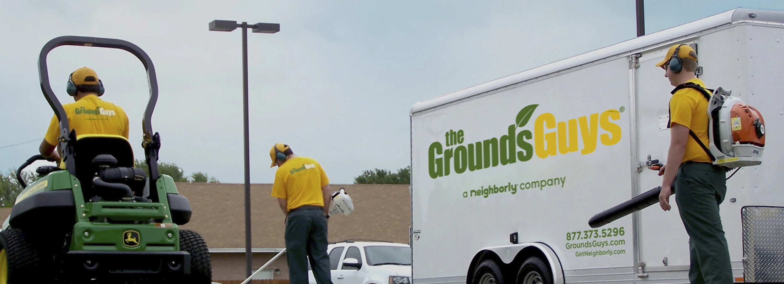 Three Grounds Guys employees performing lawn cleanup next to white trailer displaying Grounds Guys logo.