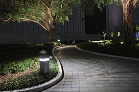 Blocky pathway lights illuminating trees outside a building and a sleek, winding walkway at night.