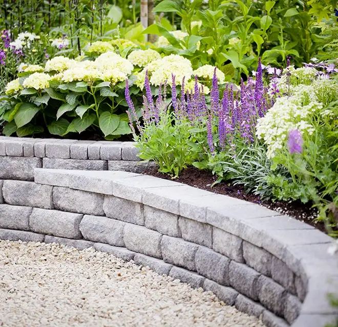 A raised plant bed with lush flowers and a stone-block retaining wall, framing a gravel path.