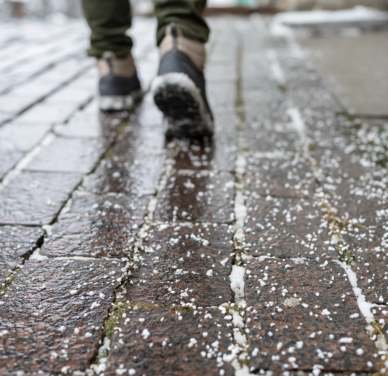 Rock salt spread on a pathway's brick pavers, with a person's boots visible walking on path.