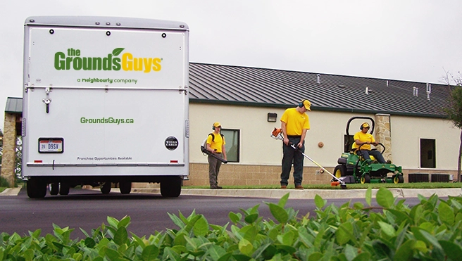 A team of The Grounds Guys service professionals cleaning up a business's lawn next to a branded trailer.