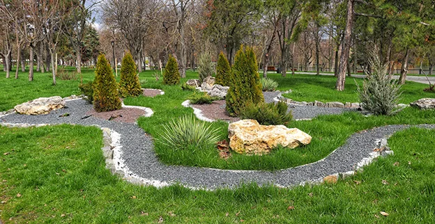 A gravel path winding around decorative rocks and shrubs, with a wooded area behind it.